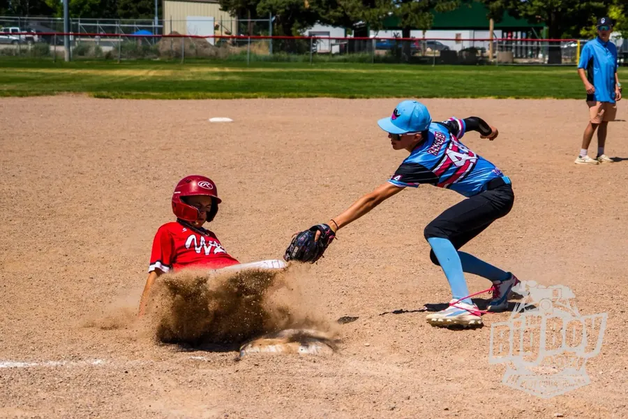 Youth baseball runner sliding into the base in a cloud of dust as the fielder applies the tag during a summer tournament game in Sidney, Nebraska